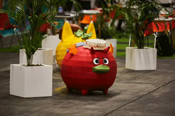 Una figura gigante de un Axie rojo entre plantas en macetas, en el lugar del evento